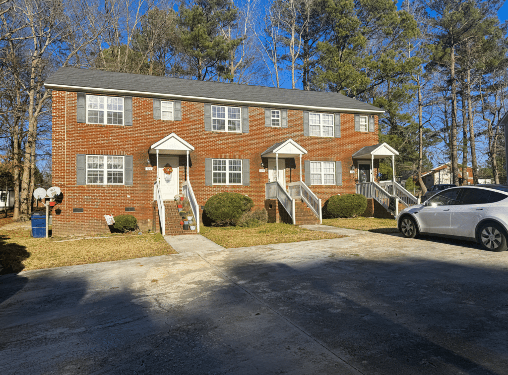 Three brick townhouses with small porches, surrounded by trees. Two satellite dishes are on the left, and a white car is parked on the right side of the driveway.